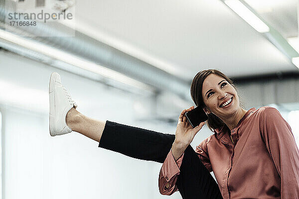 Glückliche Geschäftsfrau mit hochgelegten Beinen  die im Büro mit ihrem Smartphone telefoniert