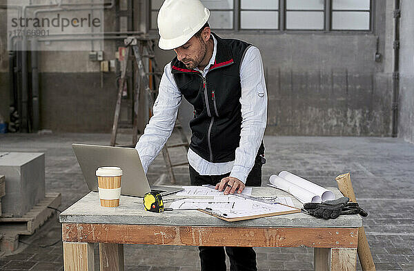 Männlicher Architekt mit Bauplänen  der einen Laptop auf einem Tisch benutzt  während er in einem Gebäude steht