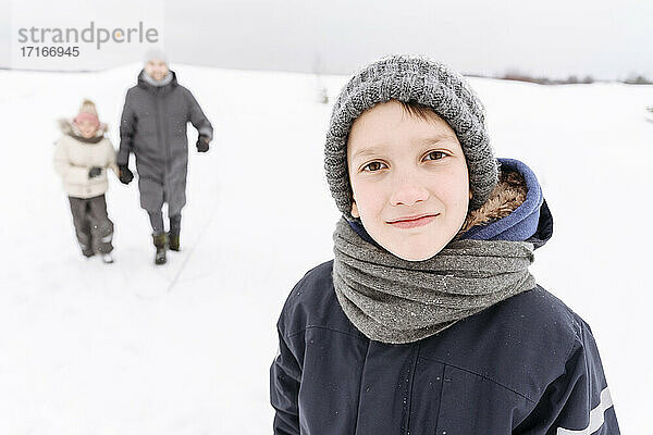 Junge  der in einer verschneiten Landschaft steht  mit Familie im Hintergrund