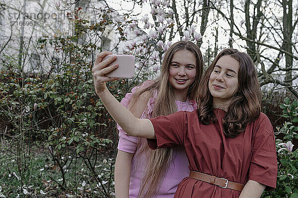 Smiling female friends taking selfie through mobile phone against magnolia tree