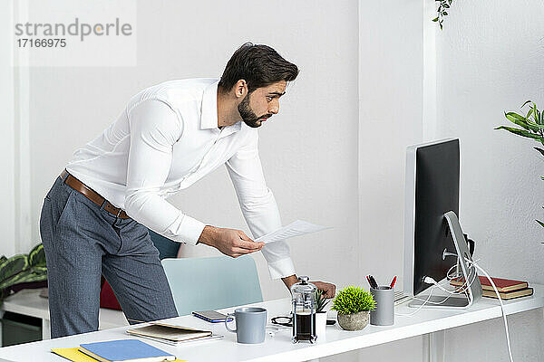 Männlicher Fachmann mit Papierdokument bei der Arbeit am Computer im Büro