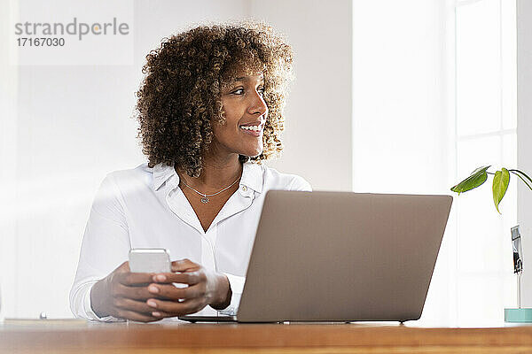 Lächelnde Geschäftsfrau mit Laptop und Mobiltelefon  die wegschaut  während sie zu Hause im Büro sitzt