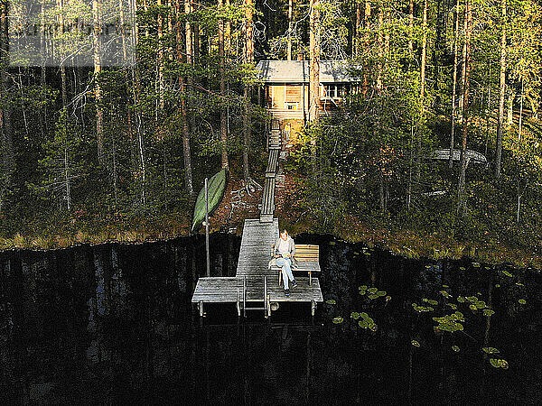 Drohnenaufnahme einer Frau  die auf einem Steg über einem See im Wald sitzt und ein Buch liest