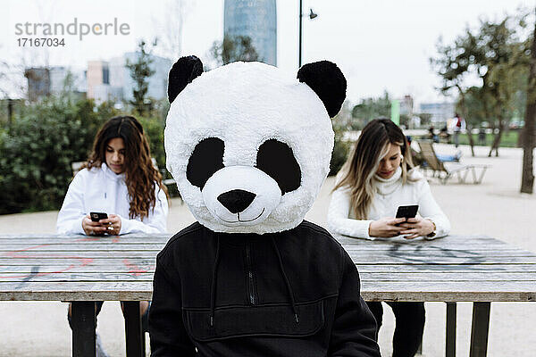 Young man with panda mask sitting against female friends at park