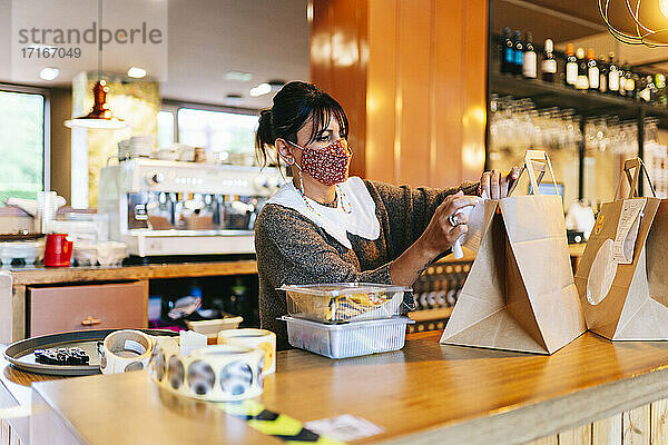Weiblicher Besitzer in Maske  der eine Quittung auf eine braune Papiertüte auf dem Tresen eines Restaurants klebt