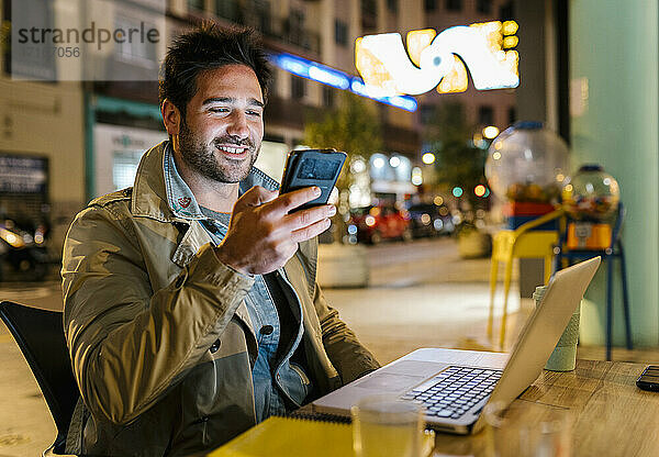 Lächelnder Mann mit Mobiltelefon  der einen Laptop benutzt  während er in einem Straßencafé sitzt