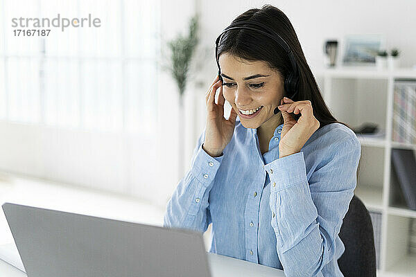 Lächelnder Kundendienstmitarbeiter beim Einstellen des Headsets während der Arbeit am Laptop im Büro