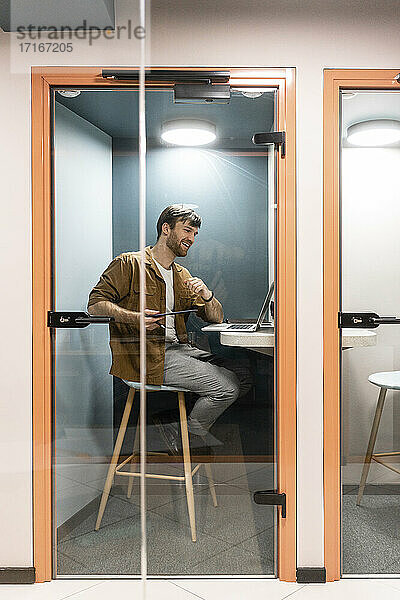 Männlicher Berufstätiger  der ein Videogespräch über einen Laptop führt  gesehen durch eine Glastür im Büro