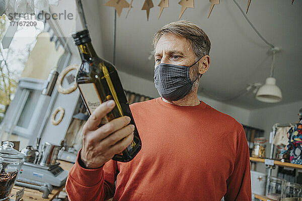 Älterer männlicher Kunde mit Maske  der beim Einkaufen in einem Einzelhandelsgeschäft eine Flasche betrachtet  während COVID-19