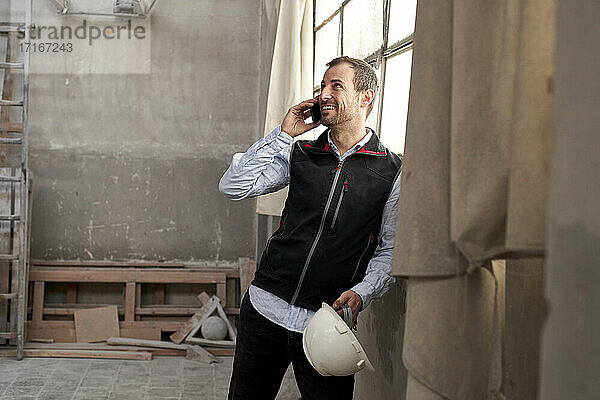 Lächelnder männlicher Bauunternehmer  der über sein Smartphone spricht  während er in einem Gebäude steht