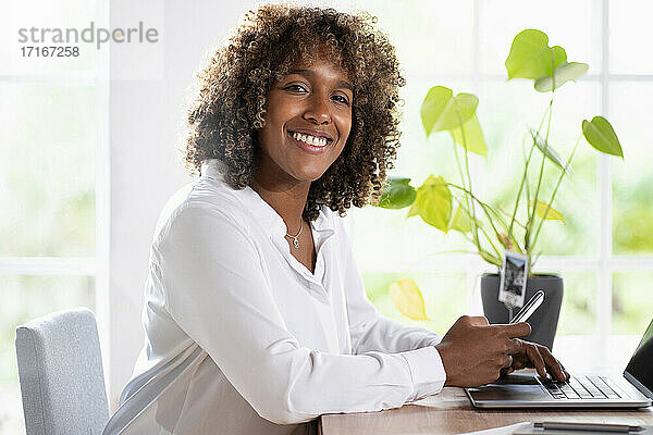 Lächelnder Unternehmer mit Laptop  der ein Mobiltelefon benutzt  während er im Büro zu Hause sitzt