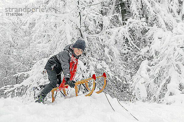 Junge beim Schlittenfahren mit Holzschlitten auf Schnee im Wald