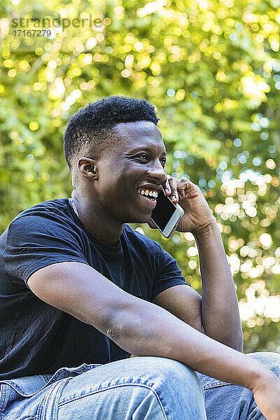Lächelnder junger Mann  der im Park sitzt und mit seinem Smartphone spricht