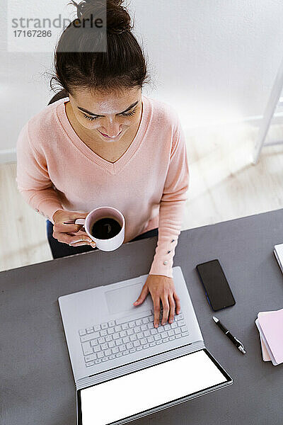 Woman with coffee cup working on laptop while sitting at home