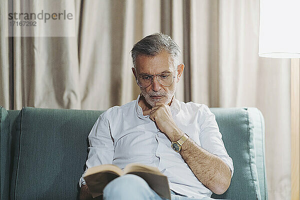 Thoughtful man with hand on chin reading book while sitting on sofa in hotel room