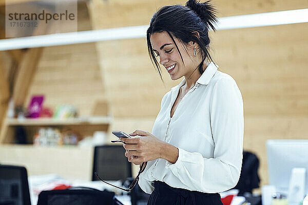 Lächelnde Unternehmerin  die im Büro eine SMS über ihr Mobiltelefon schreibt