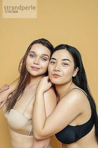 Lächelnde multiethnische weibliche Modelle in Dessous  die sich gegenseitig umarmen  während sie vor einem gelben Hintergrund stehen