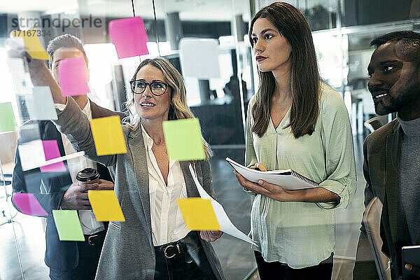 Businesswoman discussing over adhesive note on glass wall while standing with colleague in office