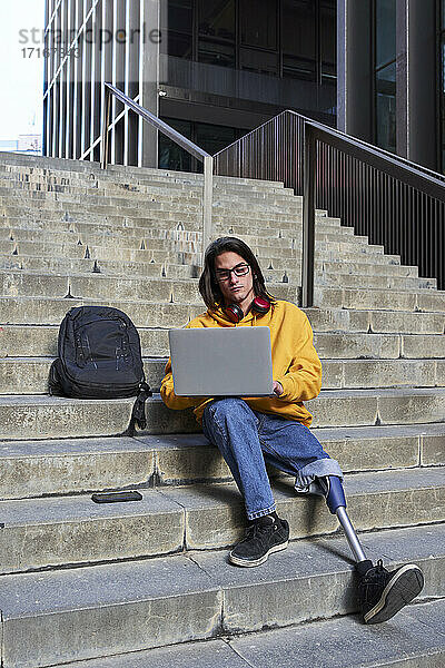 Young man with leg prosthesis using laptop while sitting on steps