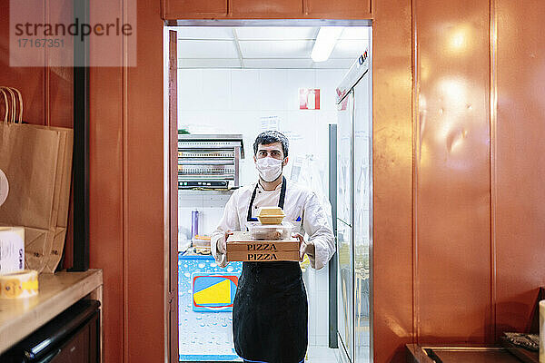 Männlicher Koch trägt Pizzakartons und Essensbehälter bei der Arbeit in einem Restaurant während einer Pandemie
