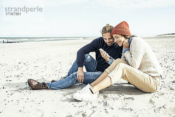 Junges Paar sitzt zusammen am Strand und benutzt ein Smartphone