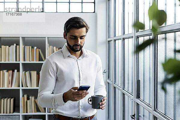 Männlicher Berufstätiger  der ein Mobiltelefon benutzt und eine Kaffeetasse im Büro hält