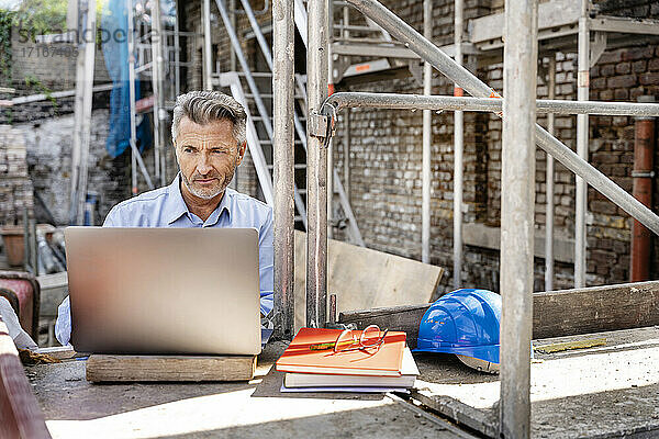 Älterer Ingenieur  der auf der Baustelle sitzend am Laptop arbeitet