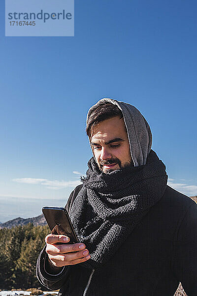 Young man in warm clothing using mobile phone against blue sky during winter
