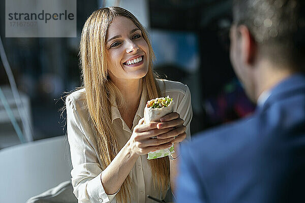 Geschäftsfrau mit Essen in der Hand und Blick auf einen in der Cafeteria sitzenden Kollegen
