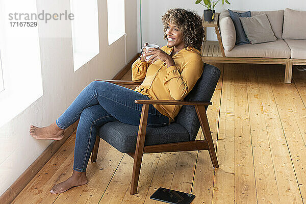 Lächelnde Frau trinkt Tee  während sie zu Hause auf einem Sessel sitzt