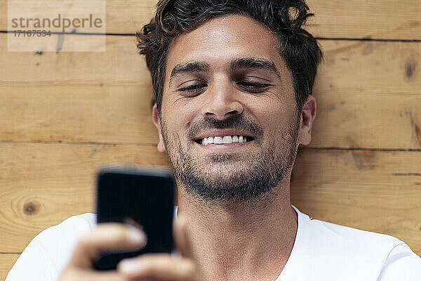 Lächelnder Mann  der ein Mobiltelefon benutzt  während er zu Hause auf dem Boden liegt