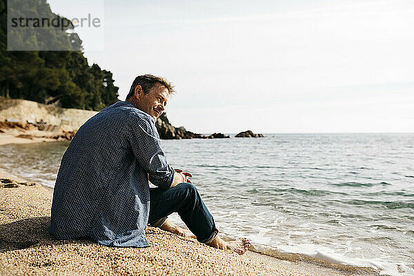 Lächelnder Mann am Strand sitzend und gegen das Meer blickend am Strand