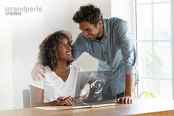Lächelndes Paar  das sich gegenseitig anschaut  während es ein Ultraschallbild auf einem transparenten Bildschirm zu Hause überprüft