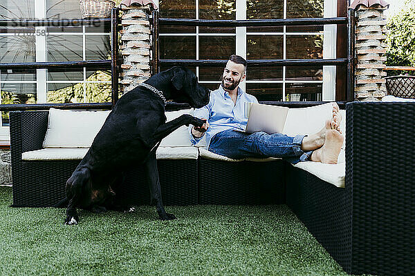 Junger Mann arbeitet am Laptop  während er mit seiner Dogge auf der Terrasse sitzt