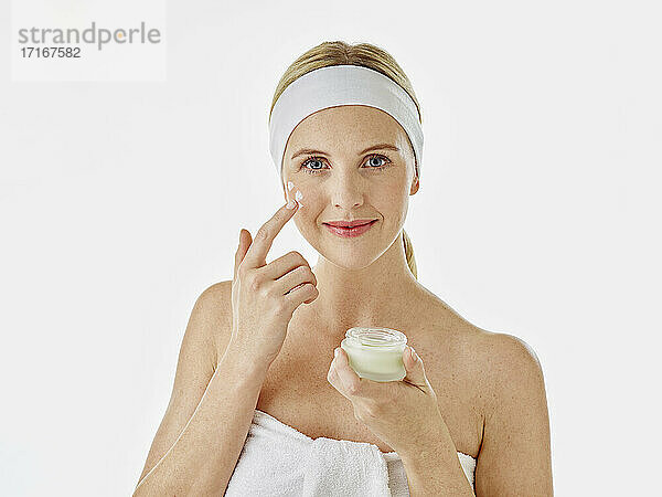 Woman wearing headband and towel applying face cream while standing against white background