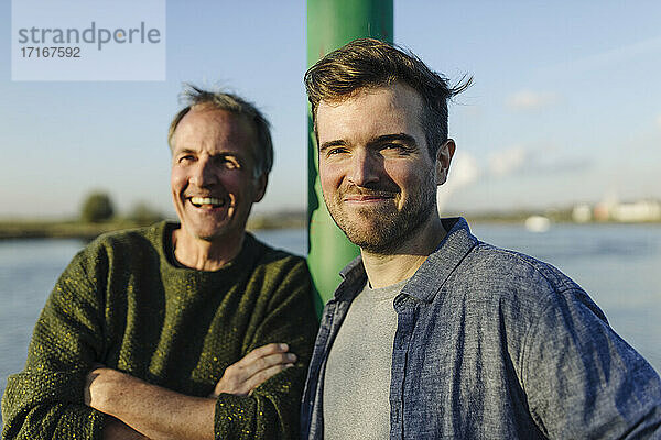 Smiling father and son by pole against river on sunny day