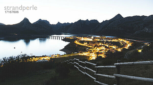 Illuminated village by lake against mountains at dusk