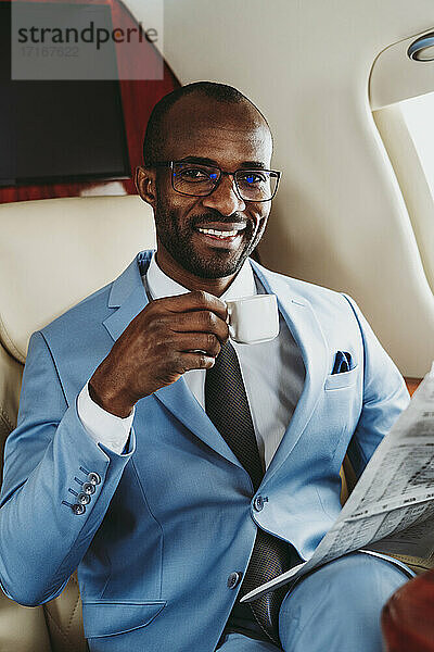Glücklicher Geschäftsmann mit Kaffeetasse und Zeitung im Flugzeug