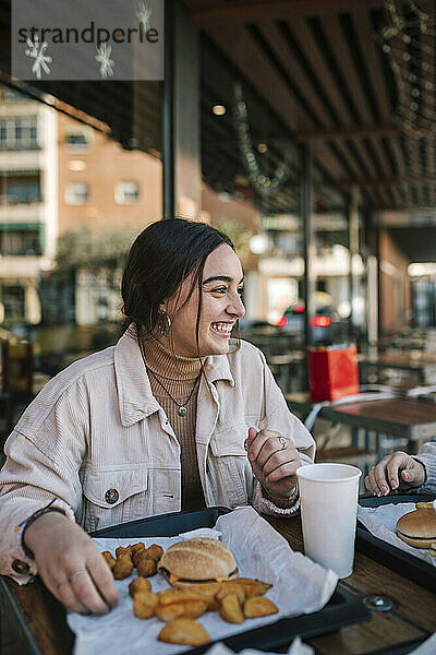 Fröhliches Teenager-Mädchen  das seinen Freund anschaut  während es in einem Restaurant im Freien Fastfood isst