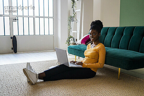 Smiling young Afro woman sitting with laptop on carpet against sofa in living room at home