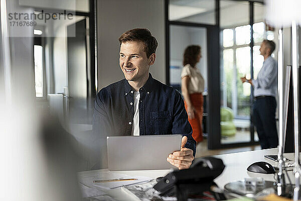 Lächelnder Geschäftsmann  der wegschaut  während er mit Kollegen im Hintergrund in einem Großraumbüro sitzt