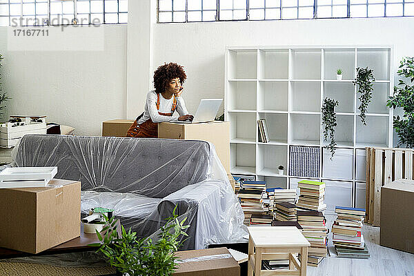 Afro-Frau mit Laptop  die sich beim Umzug in eine neue Wohnung auf einen Karton stützt
