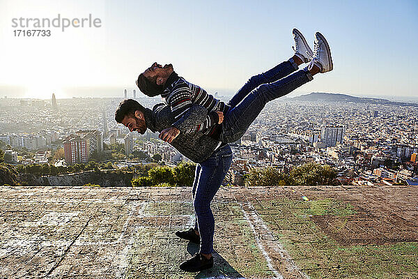 Verspielter Mann  der seinen Freund auf dem Rücken trägt  während er am Aussichtspunkt steht  Bunkers del Carmel  Barcelona  Spanien