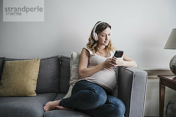Schwangere Frau hört Musik über Kopfhörer  während sie auf dem Sofa zu Hause ihr Handy benutzt