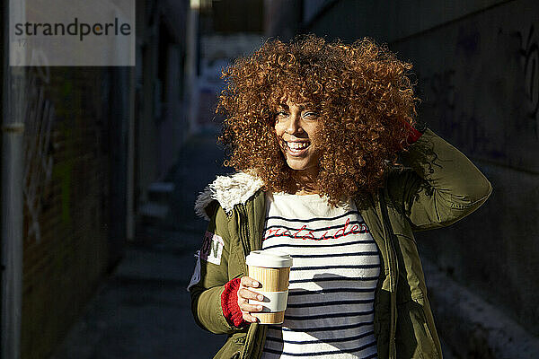 Smiling woman with hand in hair holding coffee cup while standing on footpath