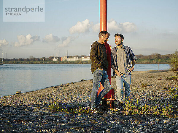 Smiling son and father leaning by pole while standing at riverbank