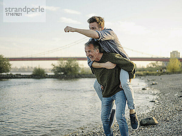 Happy father giving piggyback ride to son at riverbank against sky