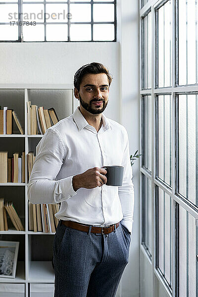 Lächelnder männlicher Unternehmer mit Händen in den Taschen  der eine Kaffeetasse im Büro hält