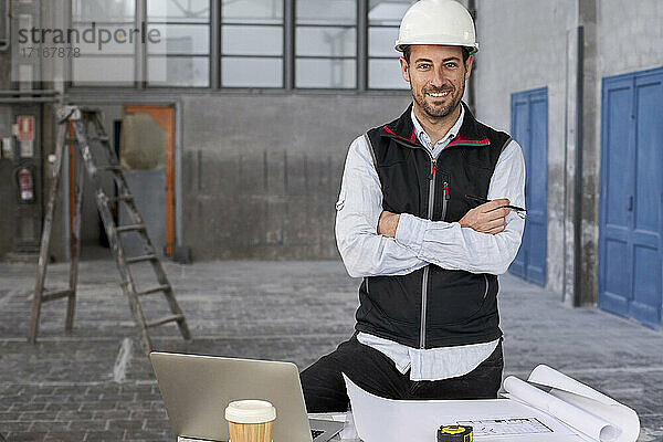 Selbstbewusster männlicher Architekt mit verschränkten Armen an einem Tisch in einem Gebäude stehend