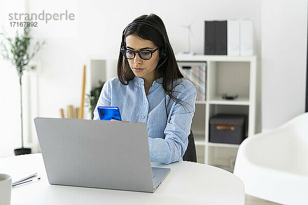 Berufstätige Frau  die ein Mobiltelefon benutzt  während sie mit einem Laptop am Schreibtisch im Büro sitzt
